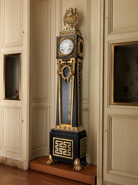 A tall dark clock case standing against panelled cream walls in a museum room. Ebony and brass, with gilt fretwork, a lion's-paw base and an urn on top. A white enamel dial with Roman numerals sits in the upper box; below it a glazed door shows the long pendulum rod hanging in the trunk.