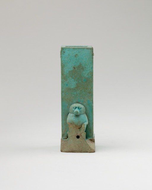A small upright vessel of turquoise-glazed faience, open at the top, with a squatting baboon modelled in relief against its lower front. A drilled hole sits at the baboon's feet, at the base of the inner chamber.