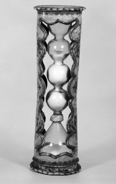 A black-and-white photograph of a tall slim hourglass in a lacquered cylindrical case, cut away on two sides so the glass shows through a scalloped opening. Inside, four bulbs of blown glass stack up to a narrow waist; the sand has run into the lower cone and the upper chamber is empty.