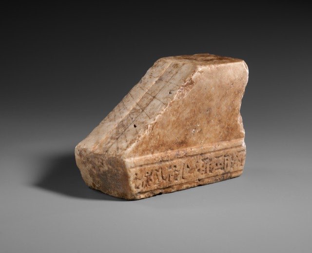 A wedge of pale weathered marble the size of two fists, photographed on grey. The sloping upper face is scored with a fan of fine straight lines running down to a rim; a band of shallow-cut hieroglyphs runs along the base of the front edge.